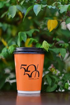 Bright orange coffee cup with logo amidst green leaves, perfect for a refreshing break.