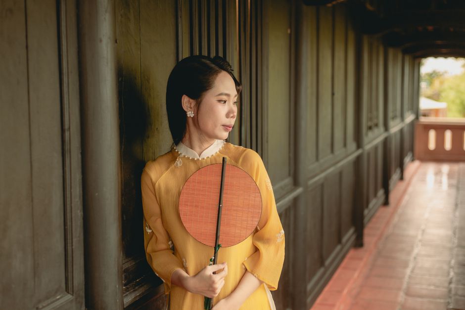 Woman in traditional yellow dress holding a fan, leaning against a wooden wall indoors.
