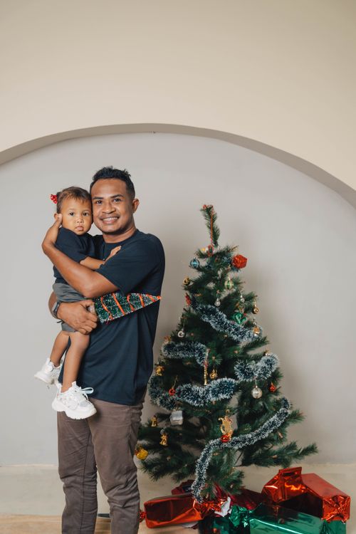 Free Father holding child beside a decorated Christmas tree indoors with gifts. Stock Photo