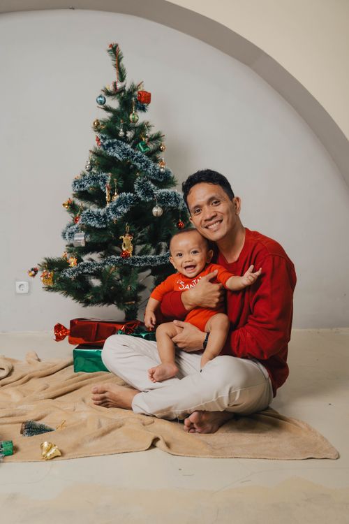 Free A joyful father and baby in festive attire celebrating Christmas by a decorated tree indoors. Stock Photo