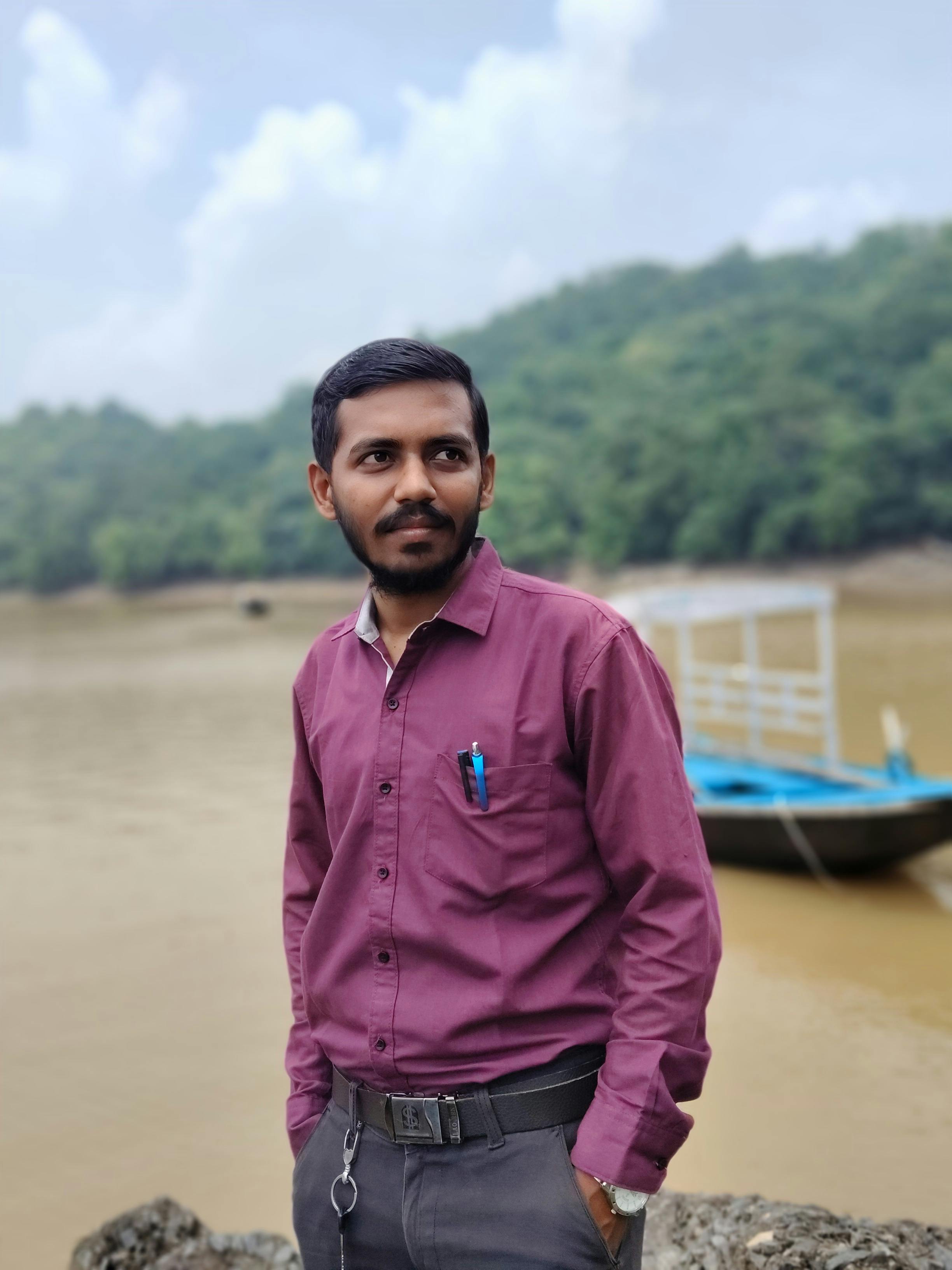 Adult man in a maroon shirt standing near a river with a boat in the background.
