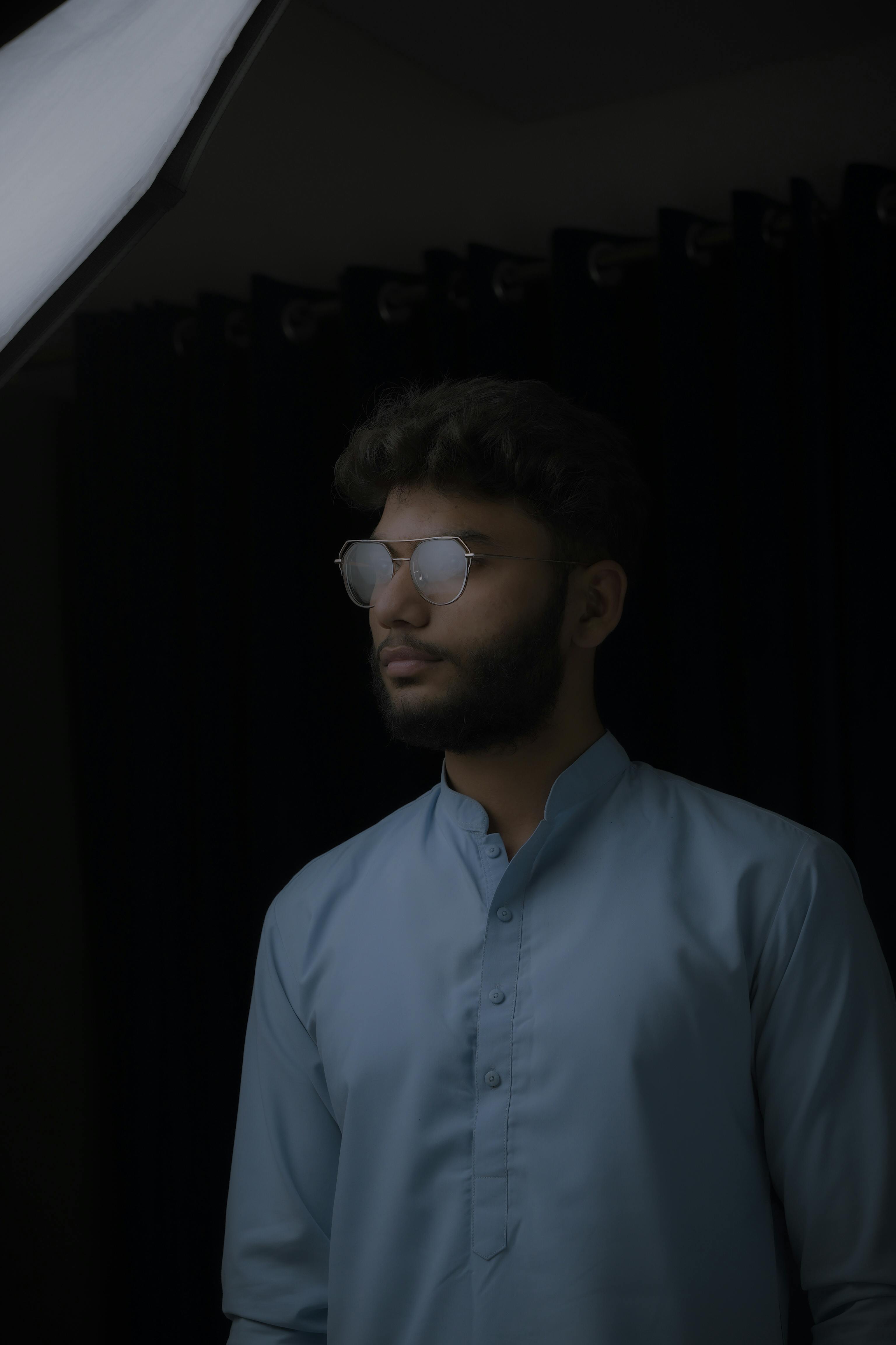 gratis Een jonge man in traditionele kleding en met een bril op, staat binnen in een peinzende houding. Stockfoto