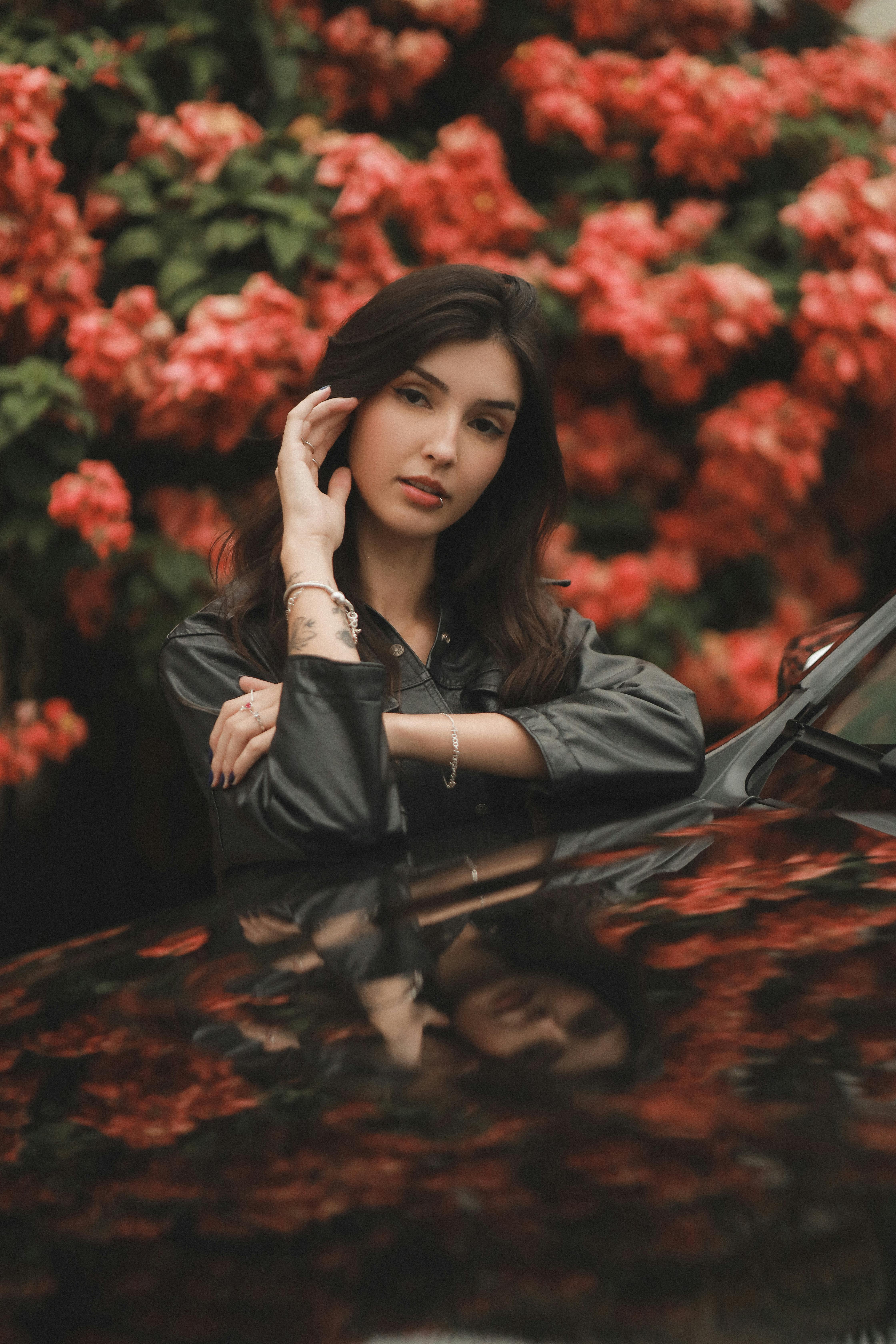 бесплатная Спокойный портрет женщины, прислонившейся к автомобилю, в окружении ярко-красных цветов. Стоковое фото