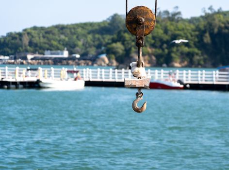 A rusted crane hook hangs over a calm waterfront with a pier, boats, and greenery in a serene summer scene.