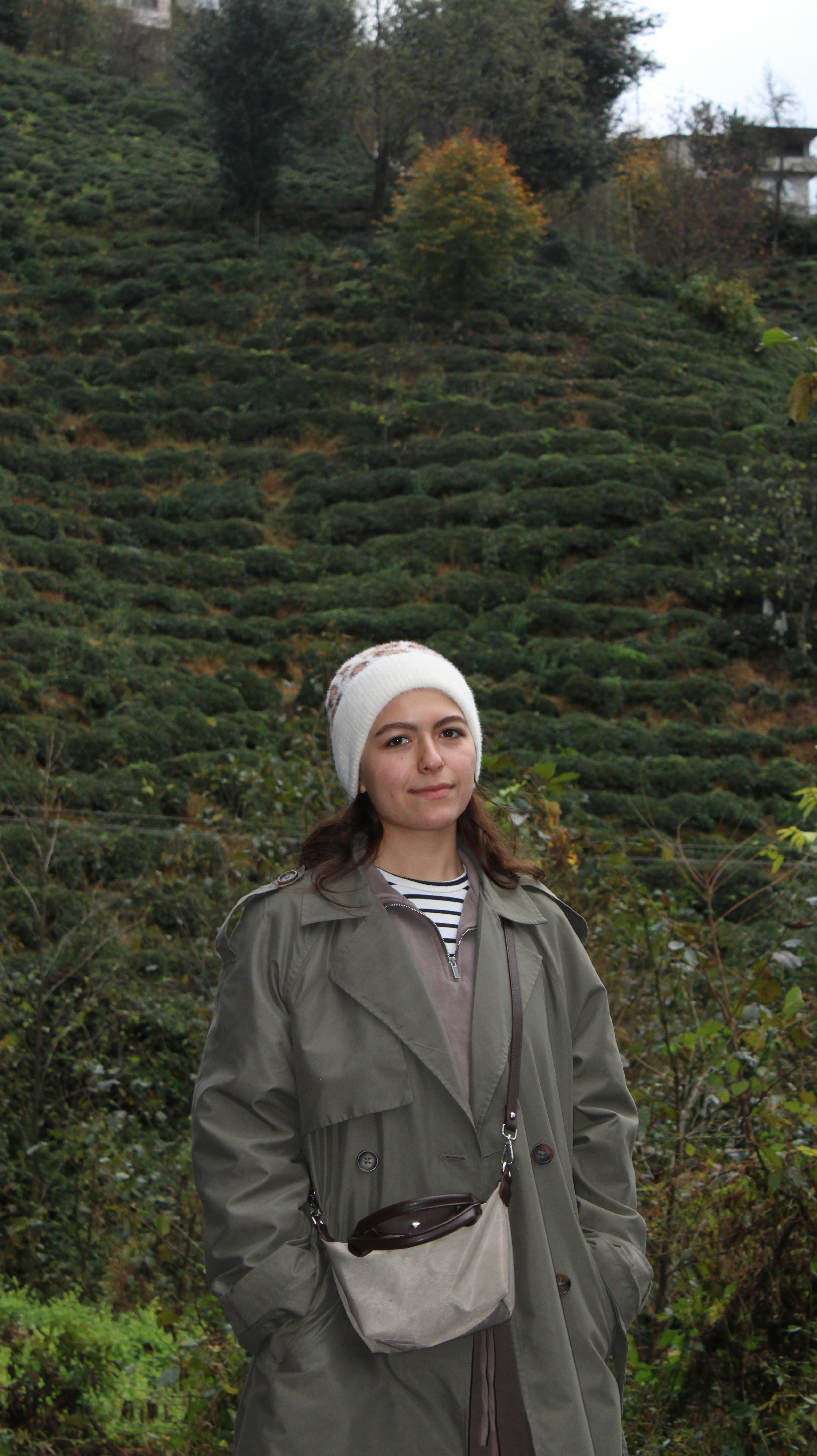Young woman dressed warmly stands in a lush, green tea plantation.