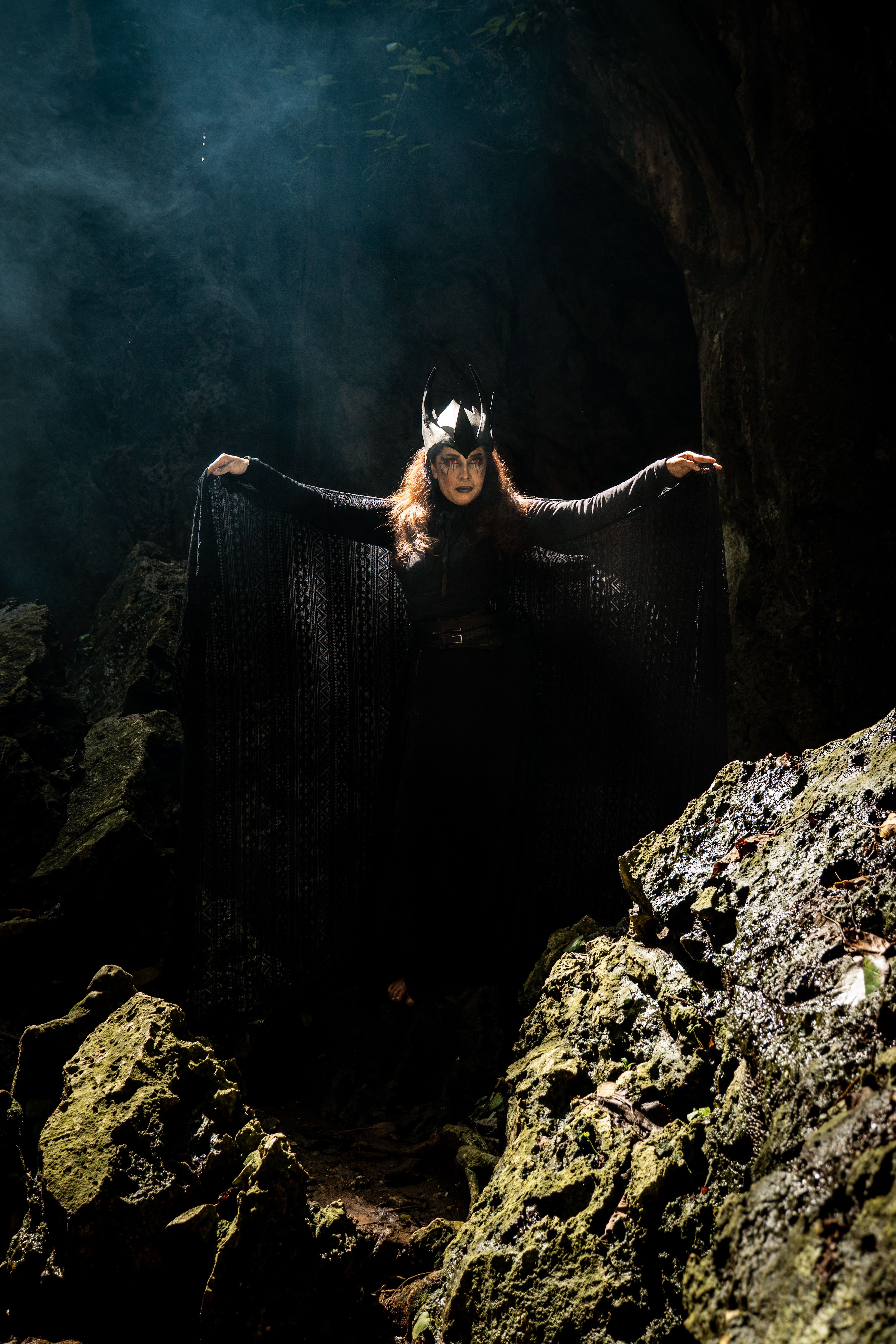 A woman in black with mystical attire stands in a forest cave, creating an enigmatic aura.