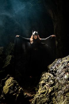 A woman in black with mystical attire stands in a forest cave, creating an enigmatic aura.