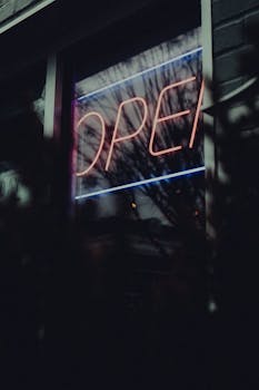 A moody urban scene featuring a neon 'open' sign reflecting in the rain outside a window.