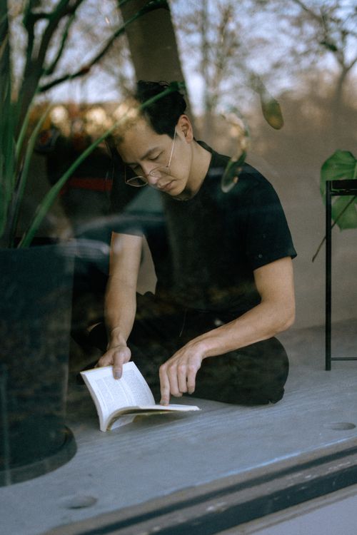 Free A young man sits by a window, engaged in reading, surrounded by indoor plants. Stock Photo