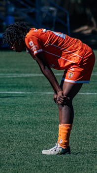 A soccer player in an orange uniform taking a break on the field during a sunny day.