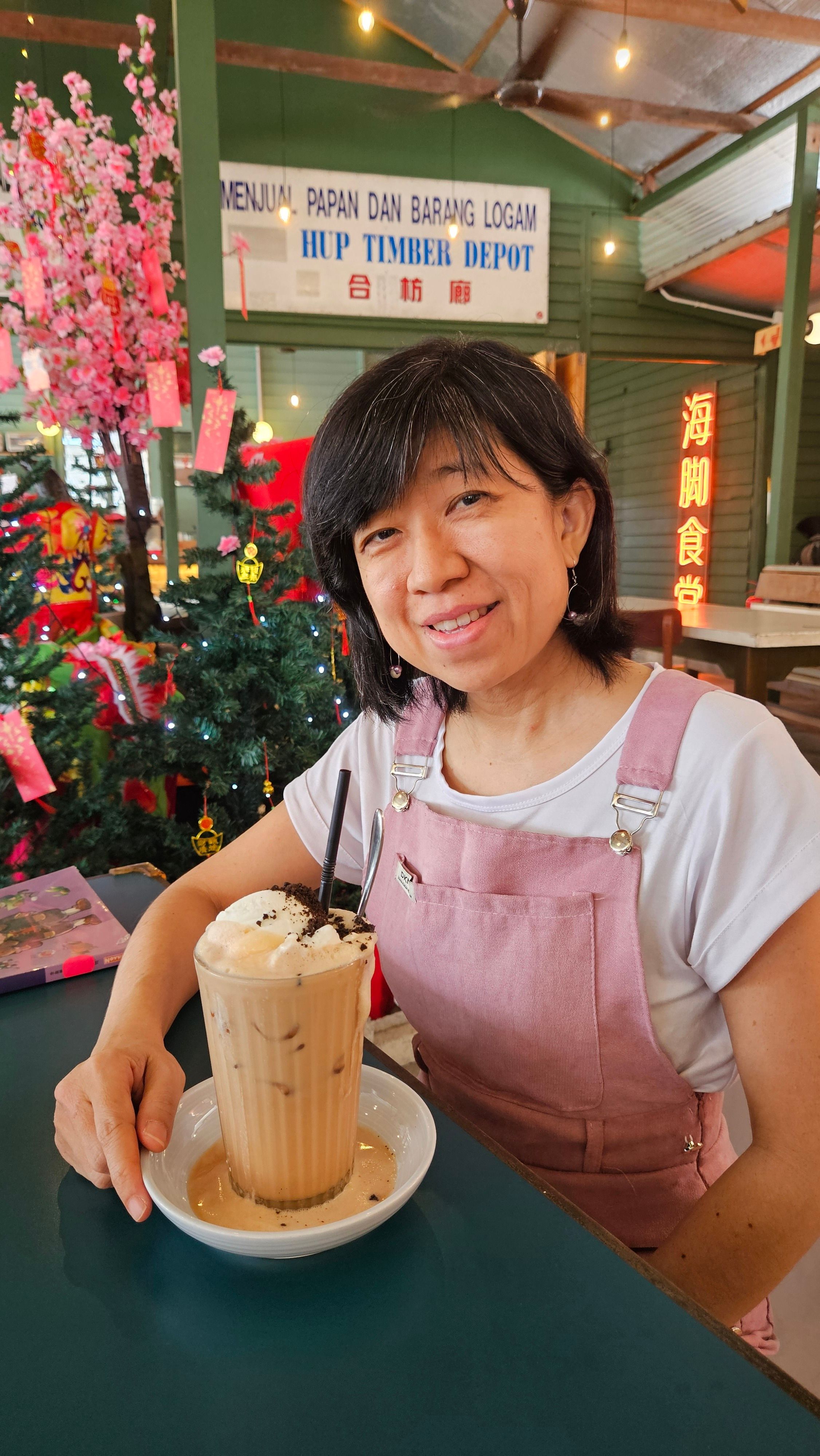 Woman in pink overalls enjoying an iced coffee indoors, featuring floral decorations.