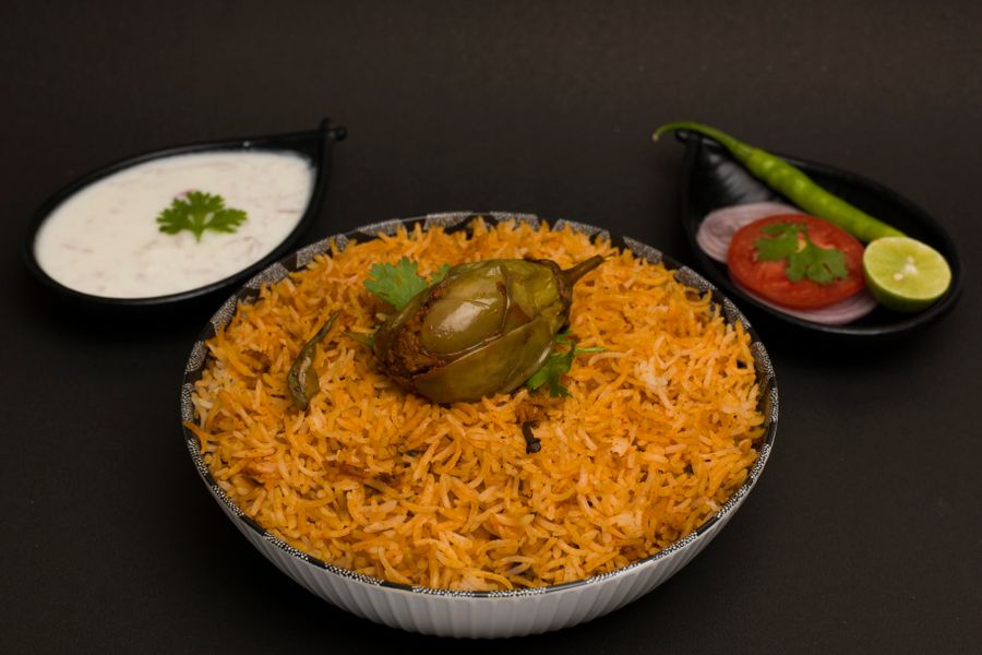 Rich Hyderabadi biryani served with raita, tomato, onion, lime, and chili garnish on a dark background.