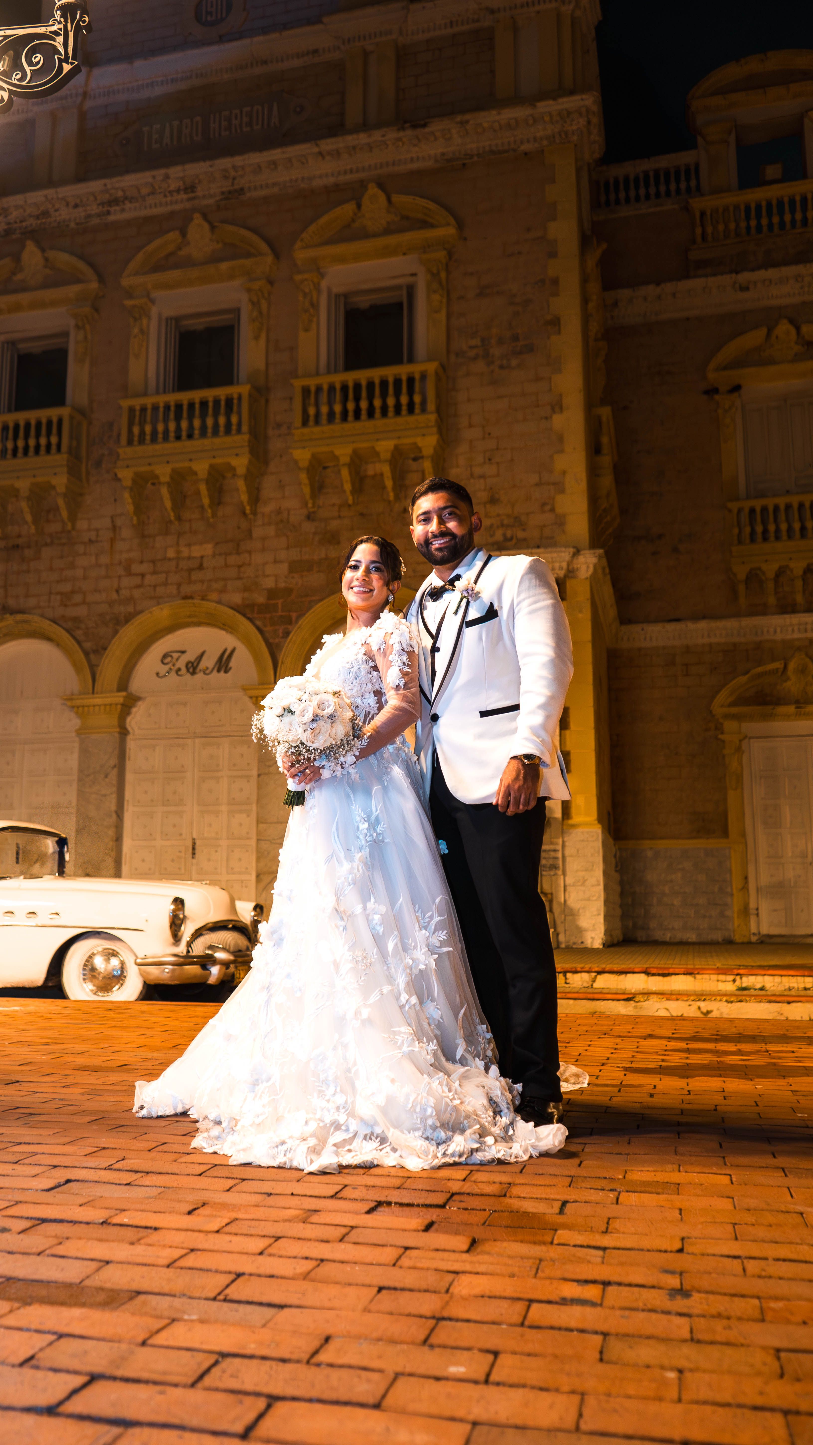 Bride and groom in elegant attire posing at night in front of an old building.