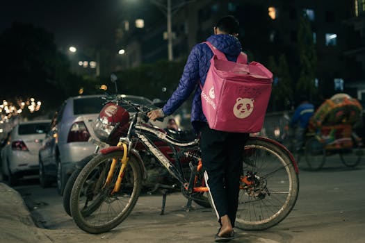A bicycle delivery person walks on a city street at night with a food delivery backpack.