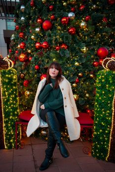 A fashionable woman poses in front of a decorated Christmas tree outdoors.