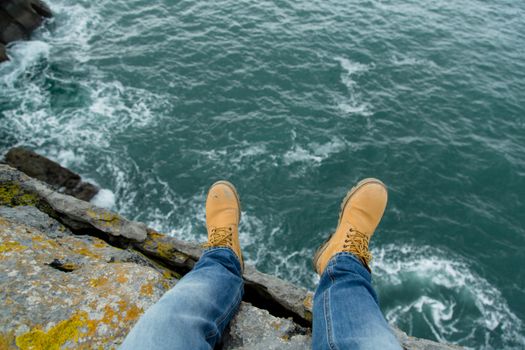 A person in boots sits on a cliff edge overlooking the ocean in Ireland, showcasing adventure and bravery.