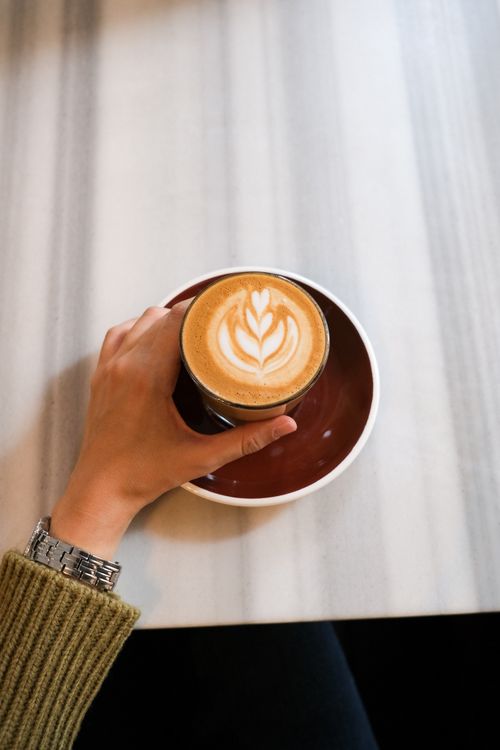Free A close-up of a hand holding a cappuccino with latte art on a cafe table. Stock Photo