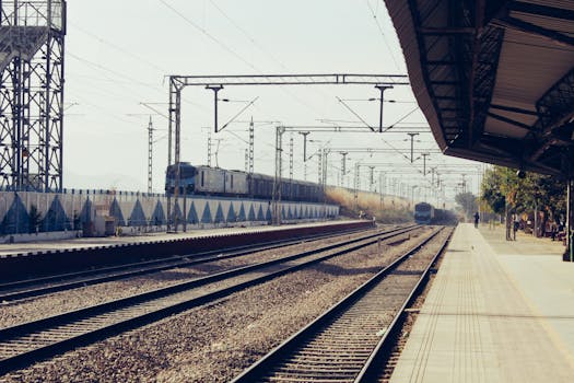Photo by Nitin Yadav A quiet railway station platform with a train passing by under the clear sky.