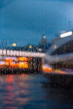 Blurred city lights seen through a rain-drenched window, creating a moody urban atmosphere.