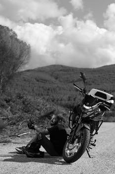Black and white photo of a motorcyclist resting beside their bike on a quiet mountain road.