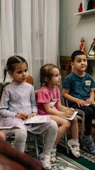 Photo by Bulat369 🌙 Three children sit attentively in a classroom setting, holding papers.