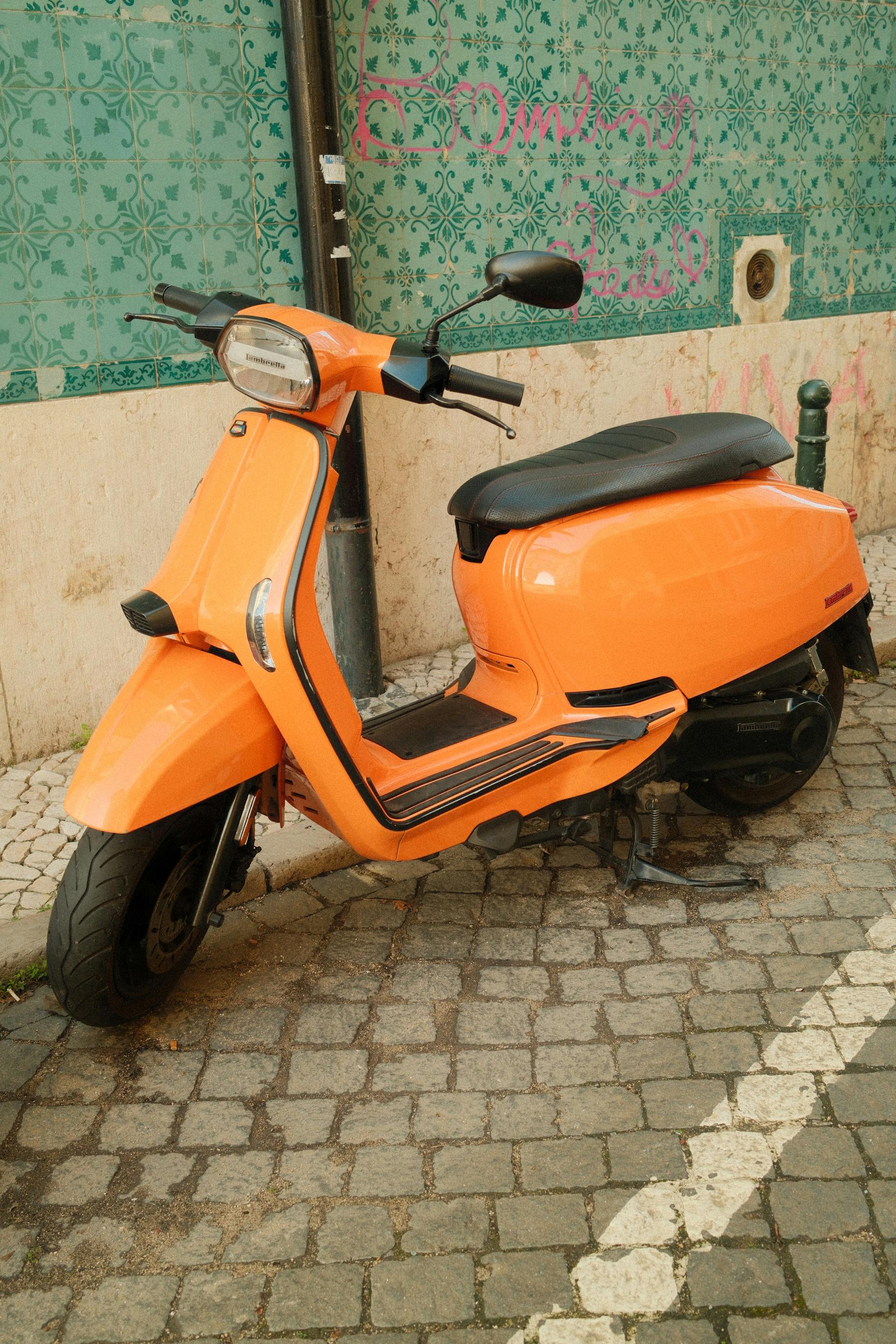 Free A vibrant orange scooter parked against a teal patterned wall on a cobblestone street. Stock Photo