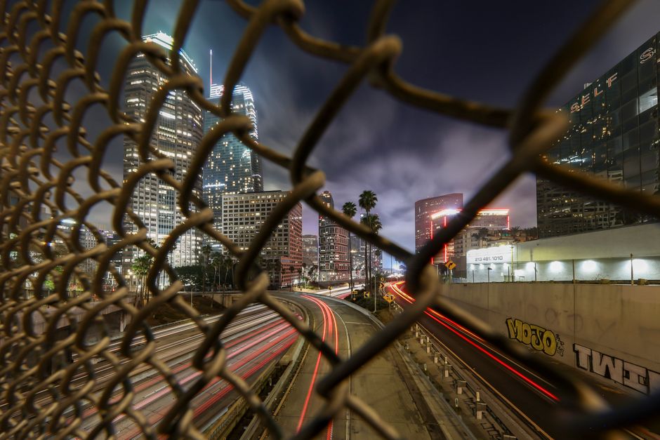 Long exposure night shot of Los Angeles cityscape through a chain-link fence with light trails.