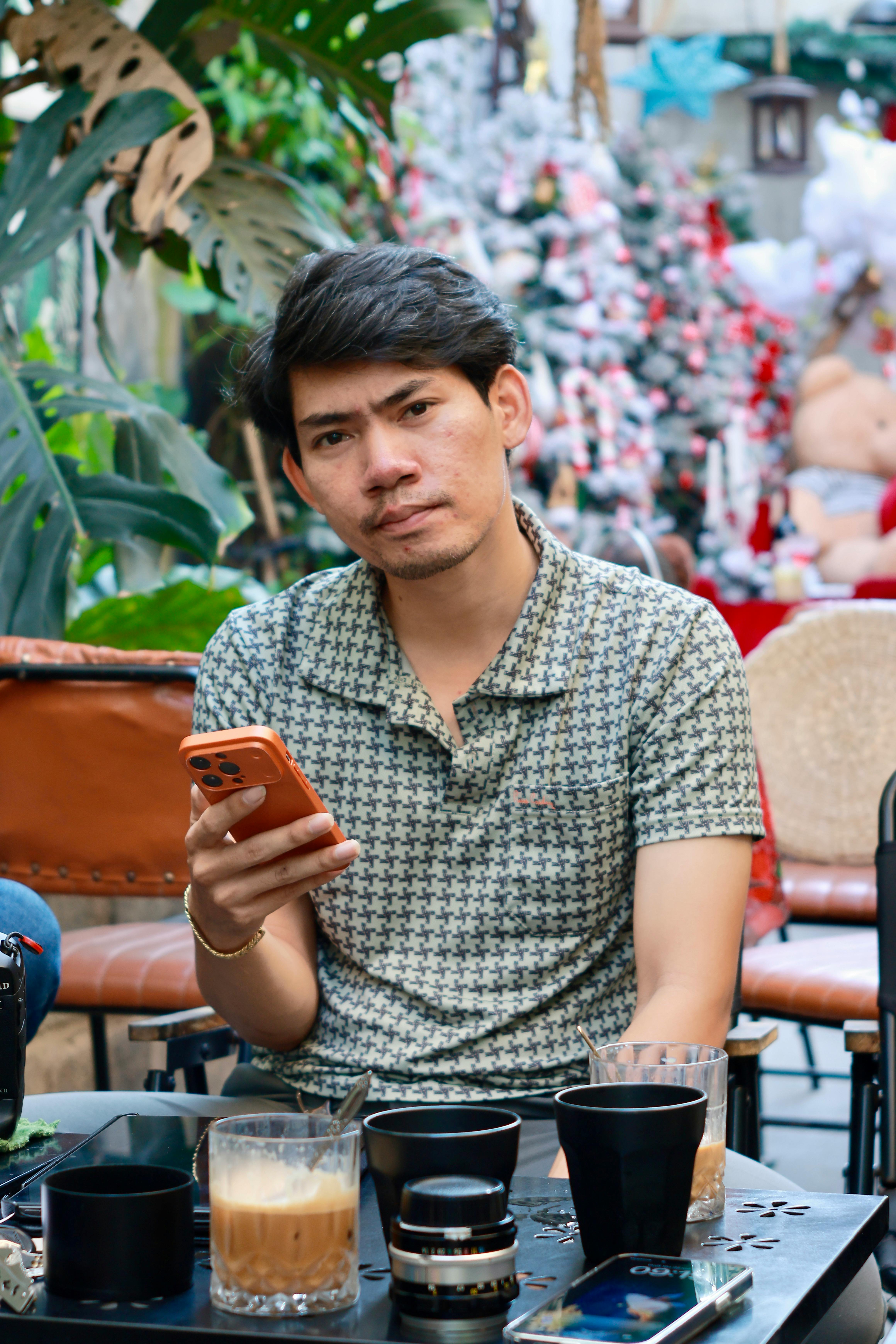 Miễn phí Chàng trai trẻ đang dùng điện thoại thư giãn tại một quán cà phê ngoài trời với không gian trang trí lễ hội. Ảnh lưu trữ