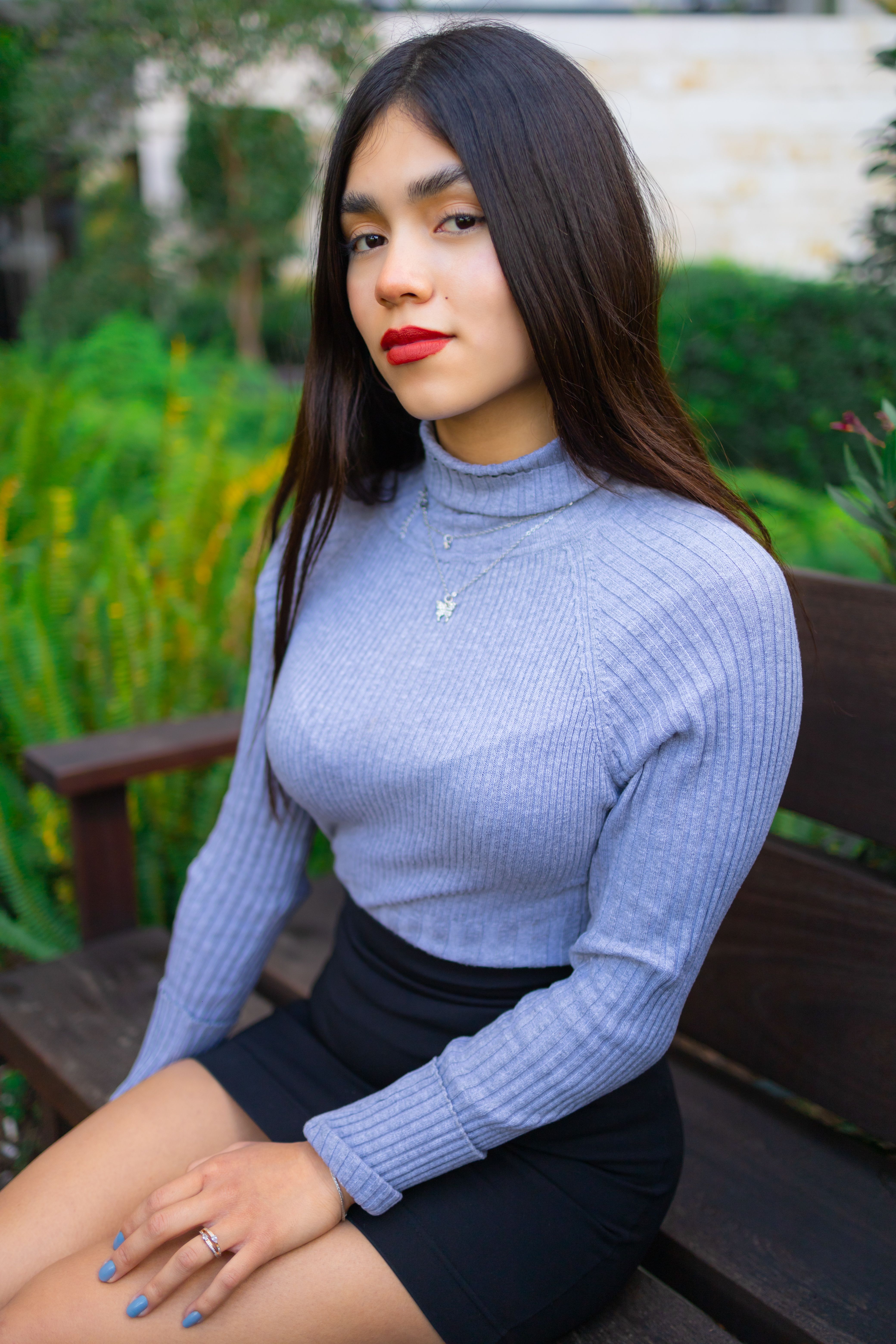 A young woman in a gray turtleneck sweater sits on a park bench with a lush green background.