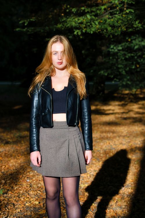 Free A woman standing in a park wearing a leather jacket during autumn sunlight. Stock Photo