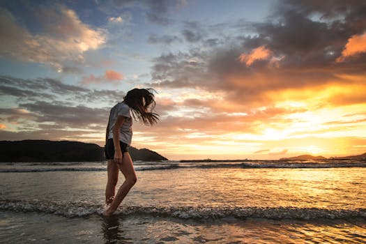 一名女子沿着海滩散步，在充满活力的日落中，海浪轻轻地拍打着她的脚