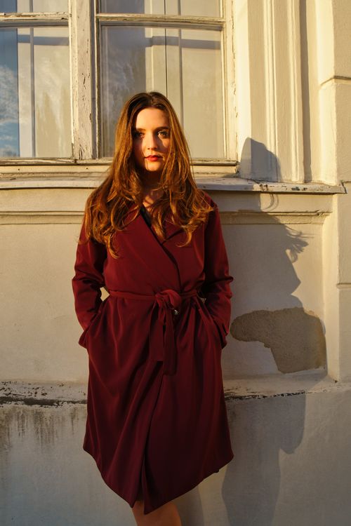 Free Young woman standing against a Berlin building at sunset wearing a burgundy coat. Stock Photo