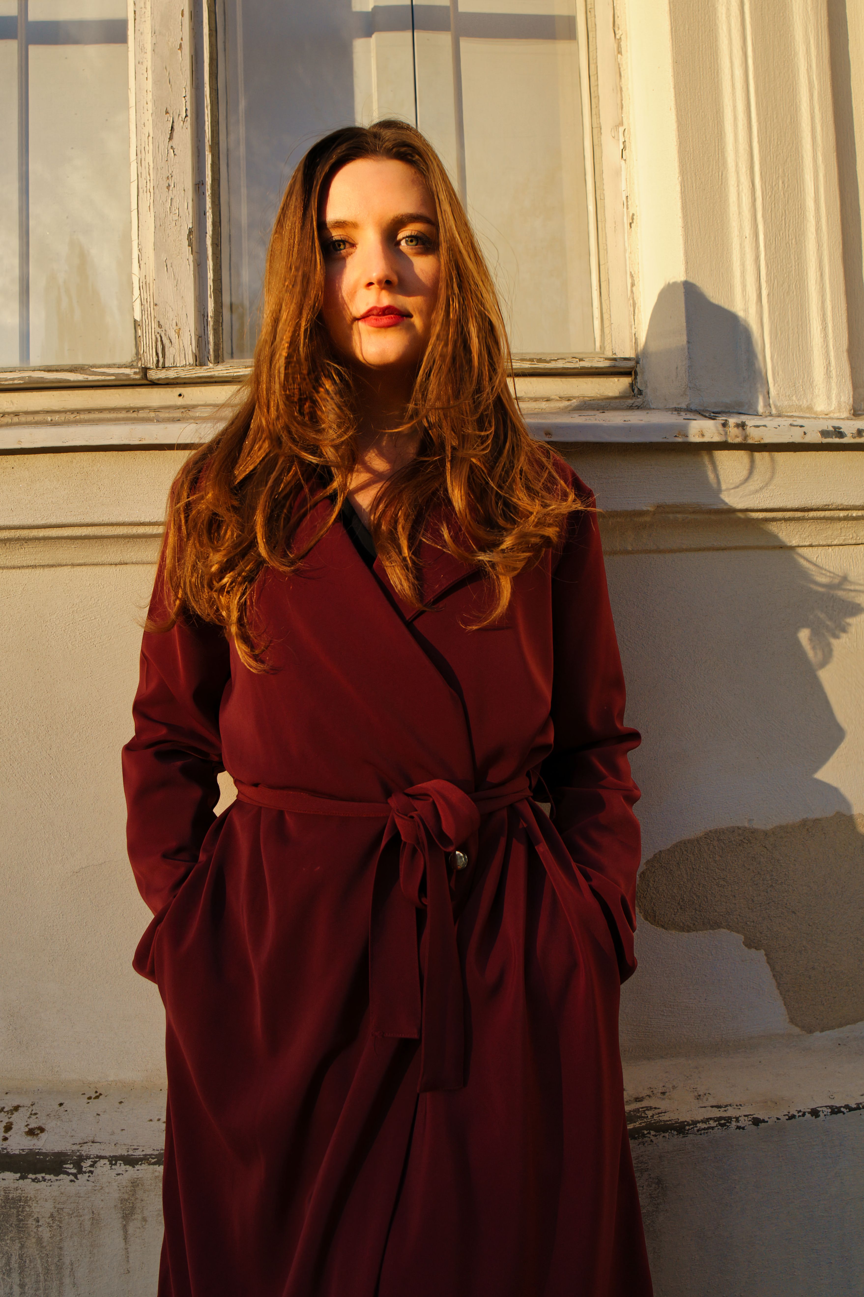 Fashionable woman in maroon coat illuminated by warm sunlight against a classic Berlin facade.