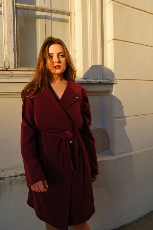 Free Fashionable woman in a burgundy coat standing by a sunlit white wall in Berlin. Stock Photo
