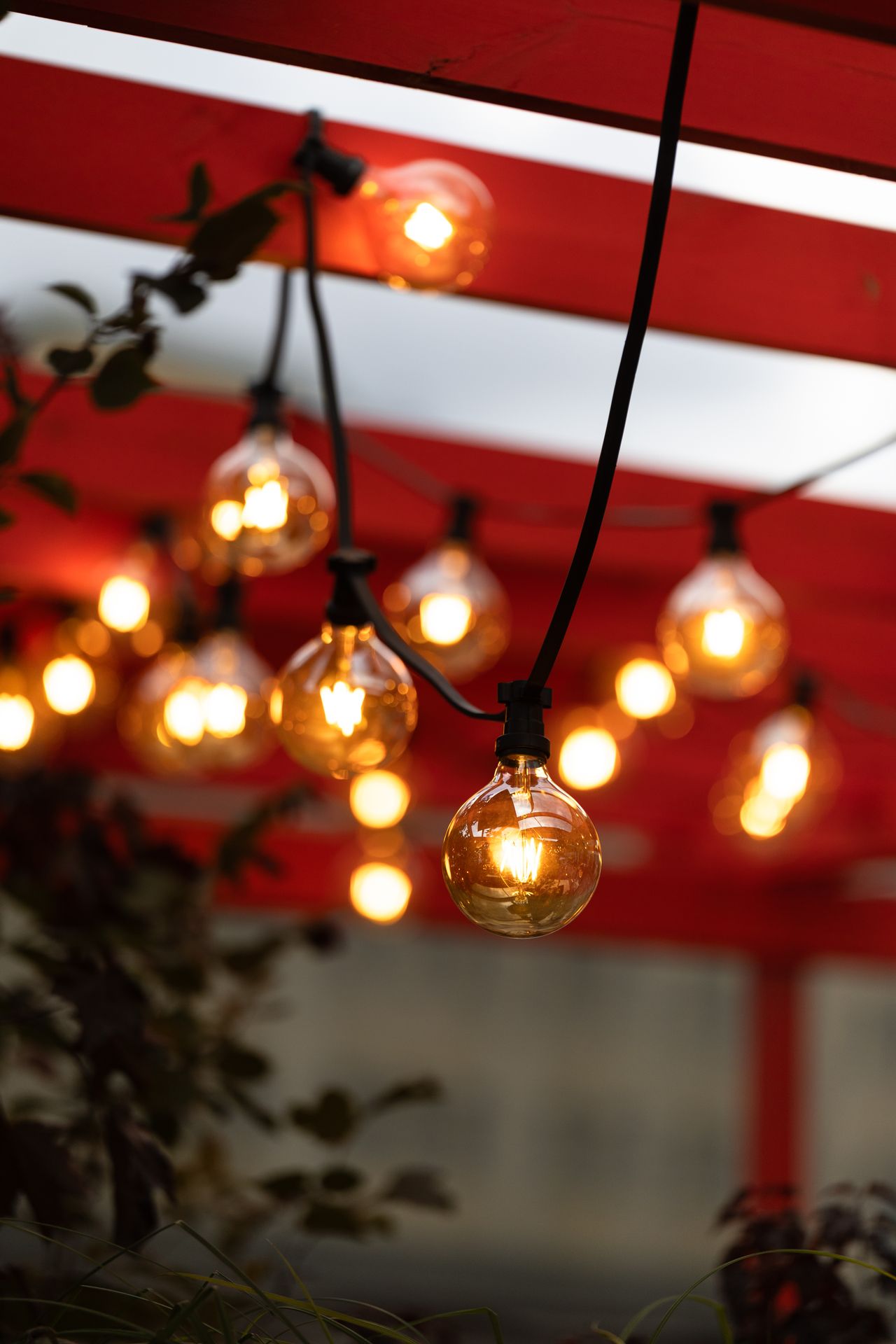 Close-up of glowing globe string light bulbs hanging from a red pergola beam in the evening