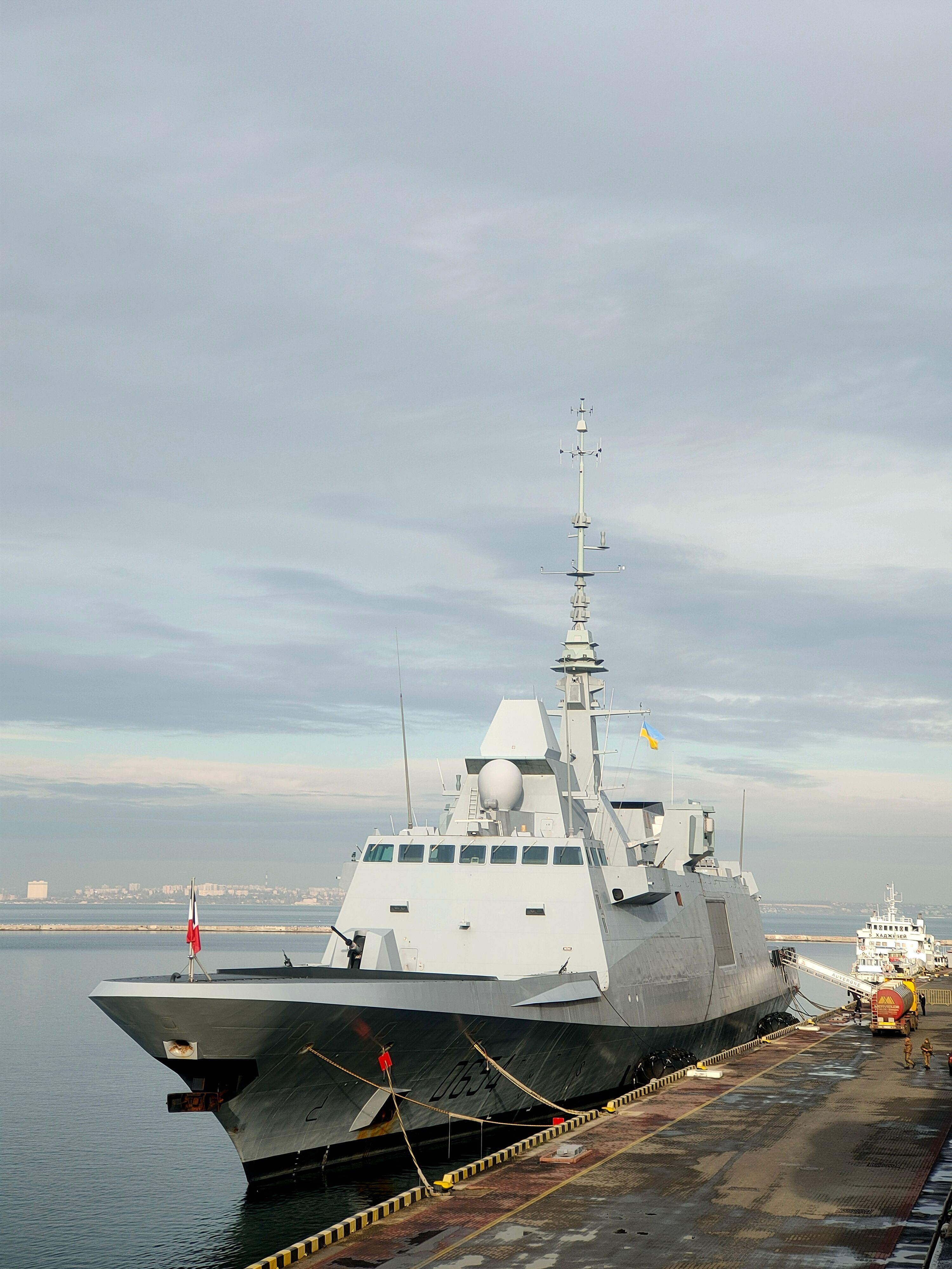 A sleek naval ship docked at a harbor under cloudy skies during the day.