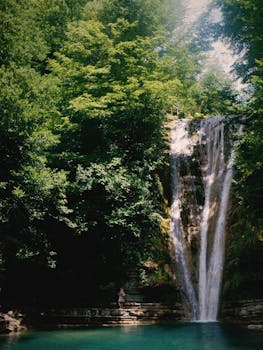 A serene waterfall cascades into a crystal-clear lake surrounded by greenery.