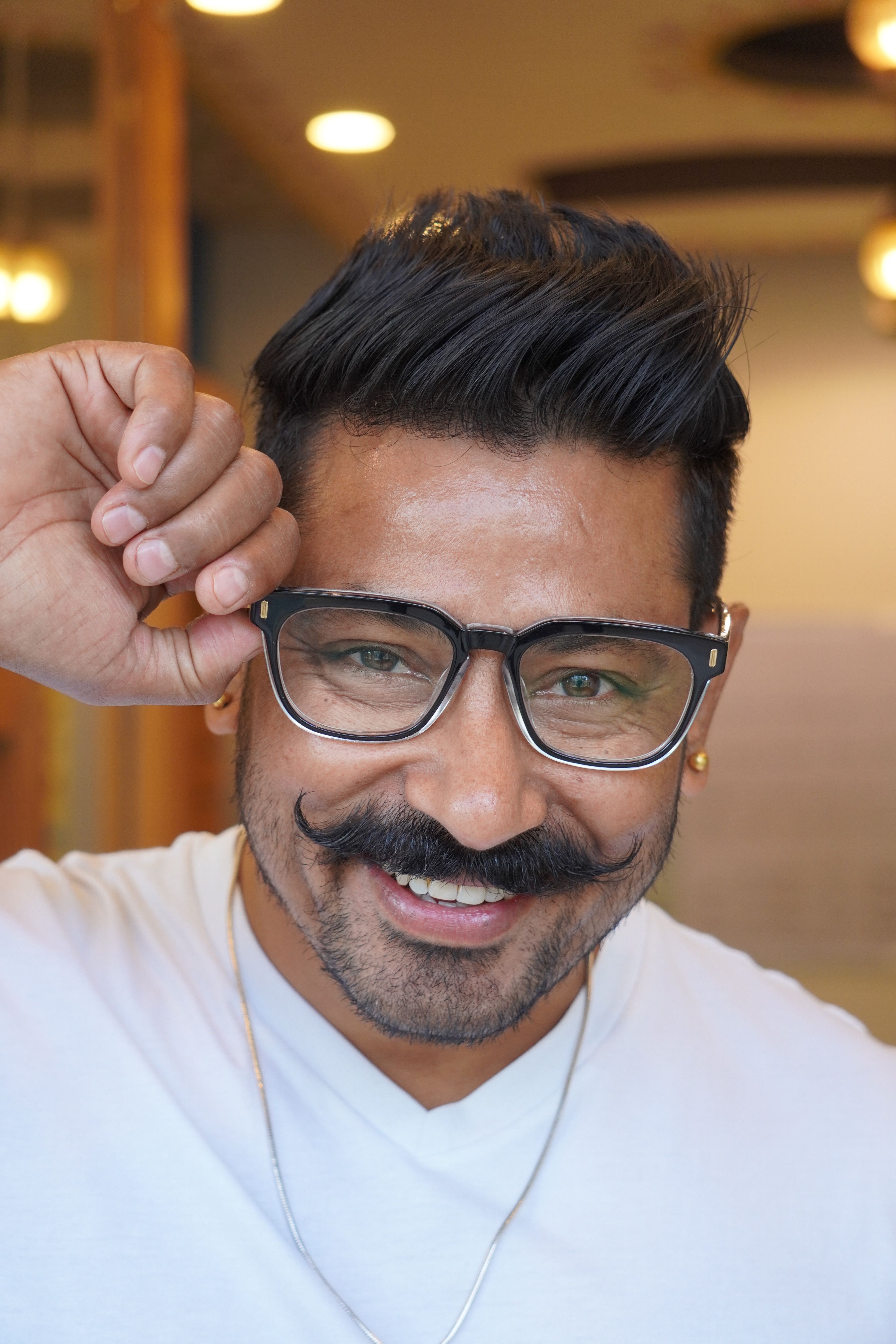 Portrait of a smiling man with glasses and a moustache in a cozy indoor setting.