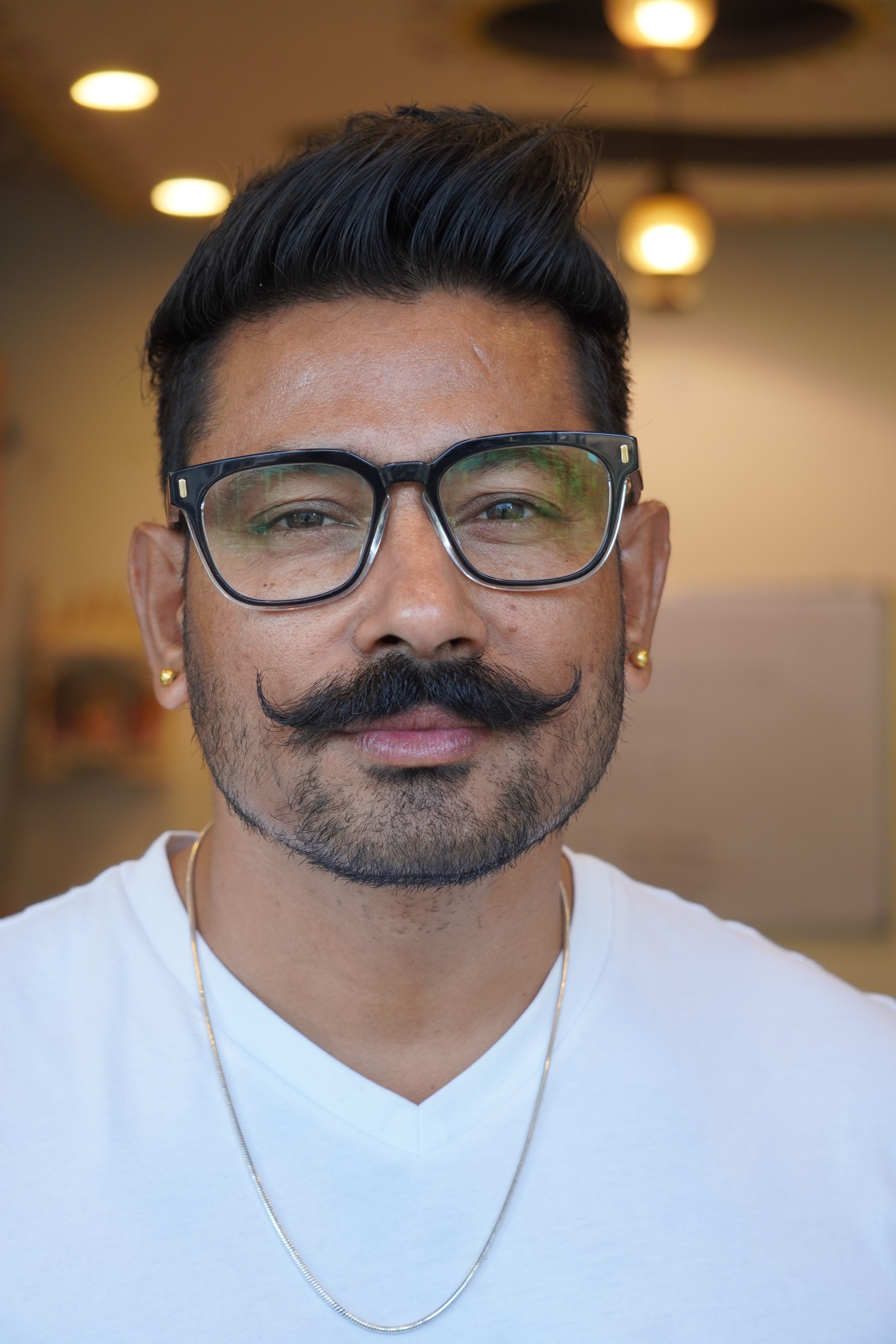 Close-up portrait of a middle-aged man with styled mustache and glasses indoors.