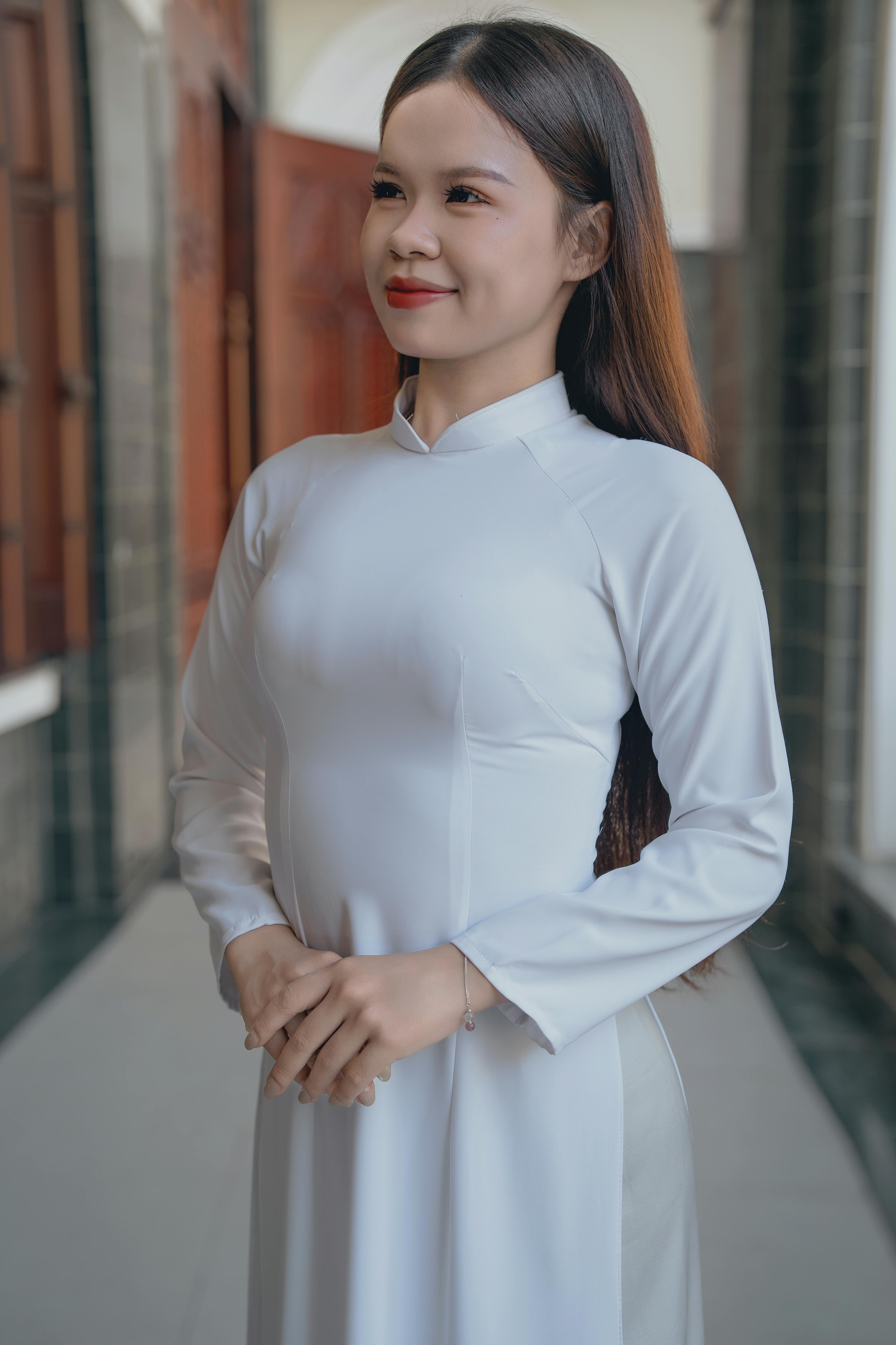 Smiling woman in a white Ao Dai standing outdoors with folded hands.