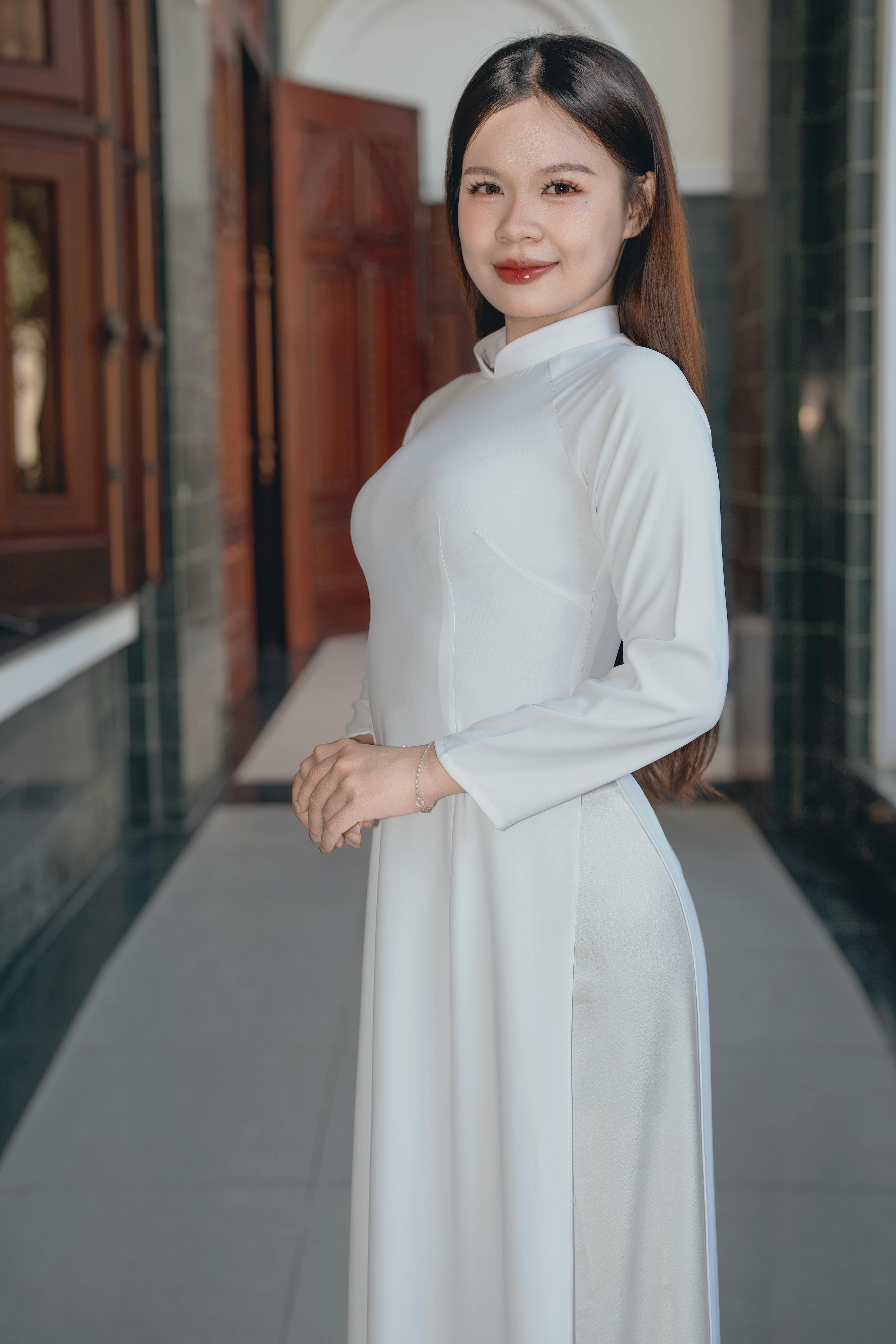 Portrait of a young woman wearing a traditional white Ao Dai, standing indoors with a warm smile.