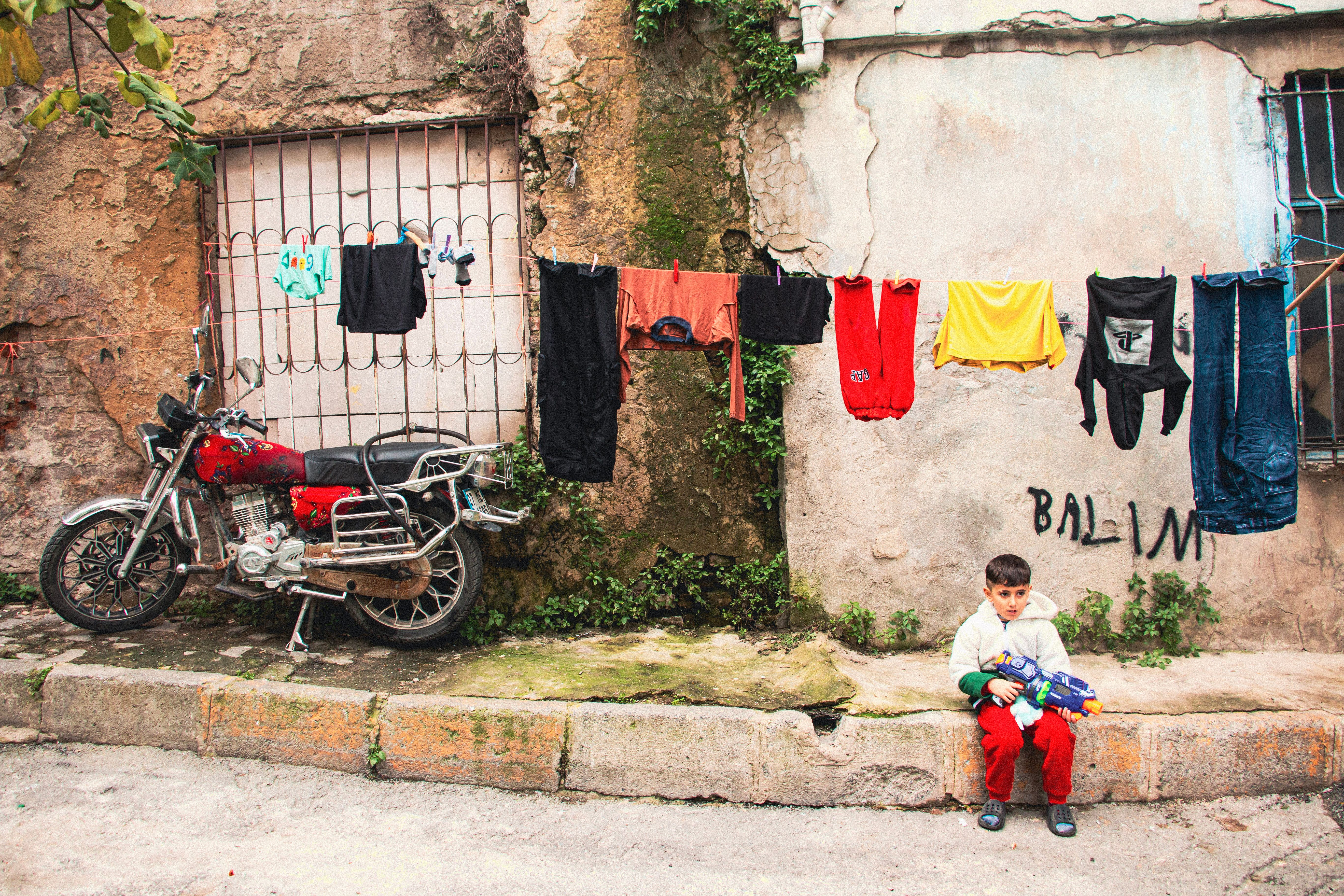 A child sits by a clothesline with a motorcycle, against a textured wall in a rustic setting.