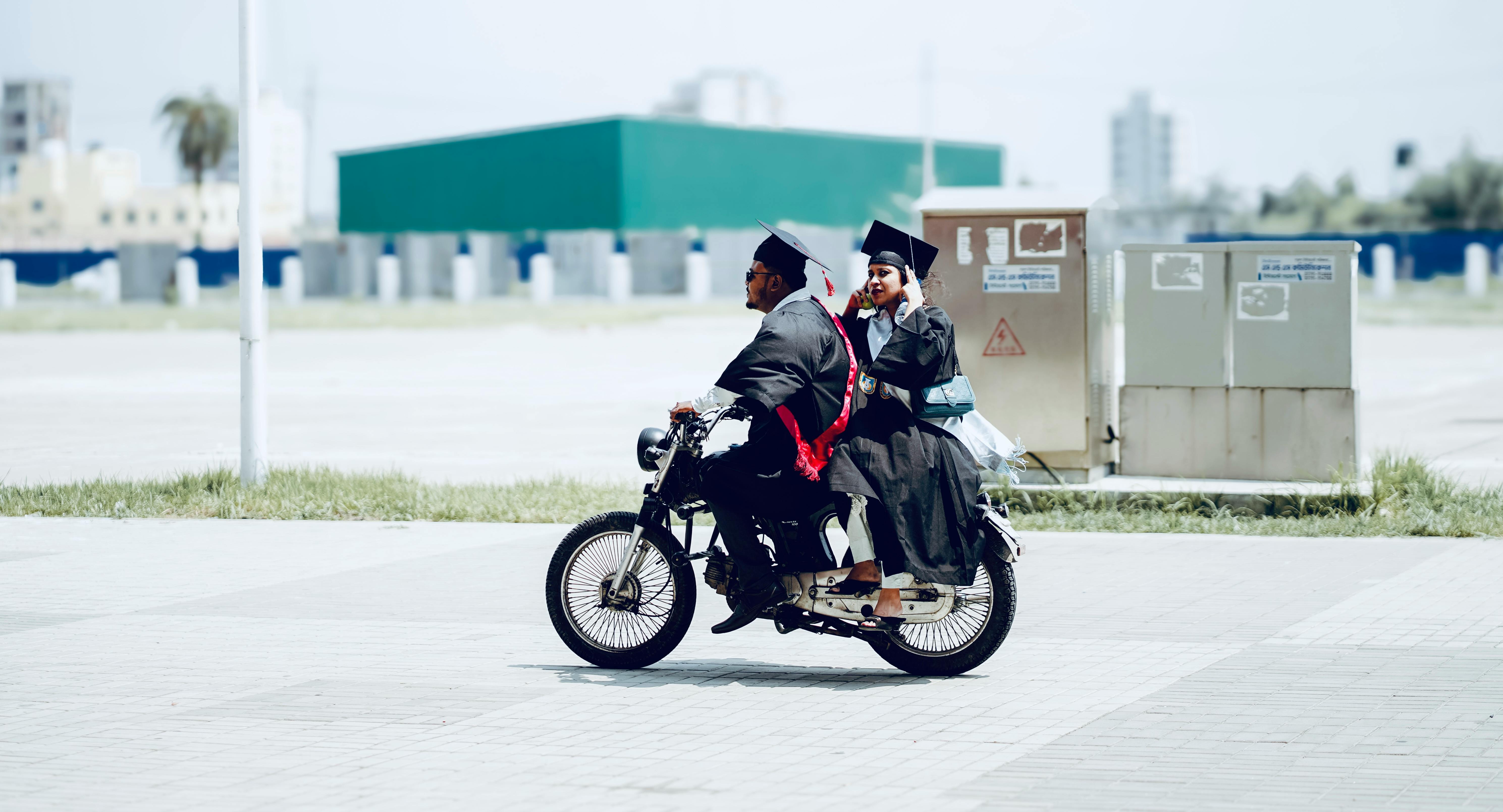 Two graduates in caps and gowns ride motorcycle outdoors celebrating.