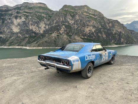 A vintage blue car parked by a scenic mountain lake with rocky cliffs.