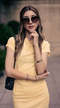 Fashionable woman in sunglasses poses confidently in a yellow dress outdoors.