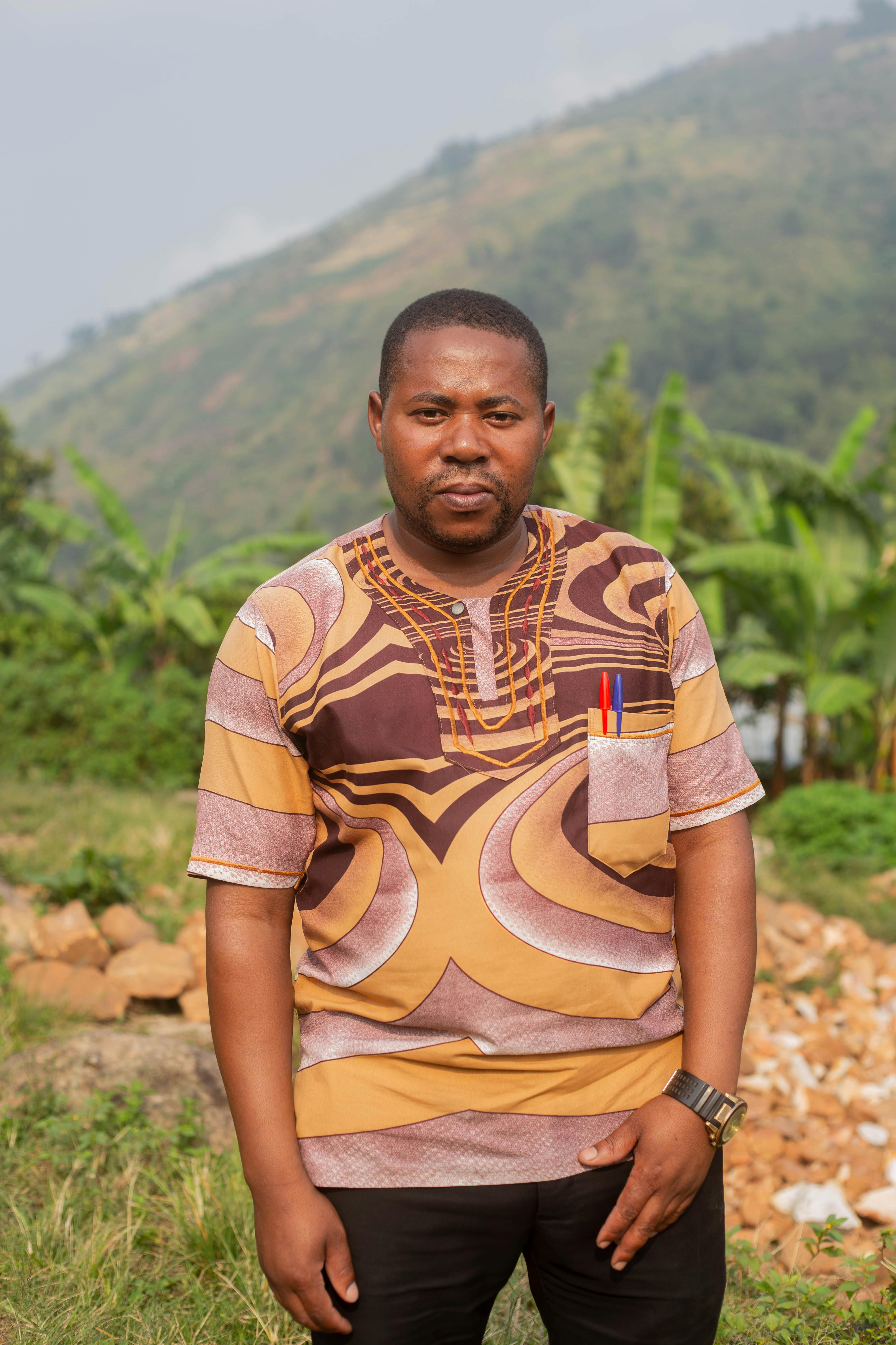 Portrait of an adult man in colorful traditional African clothing in an outdoor setting with lush greenery.