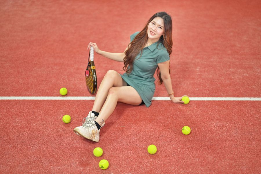 A player in a green tennis dress sits on a terracotta court among scattered balls