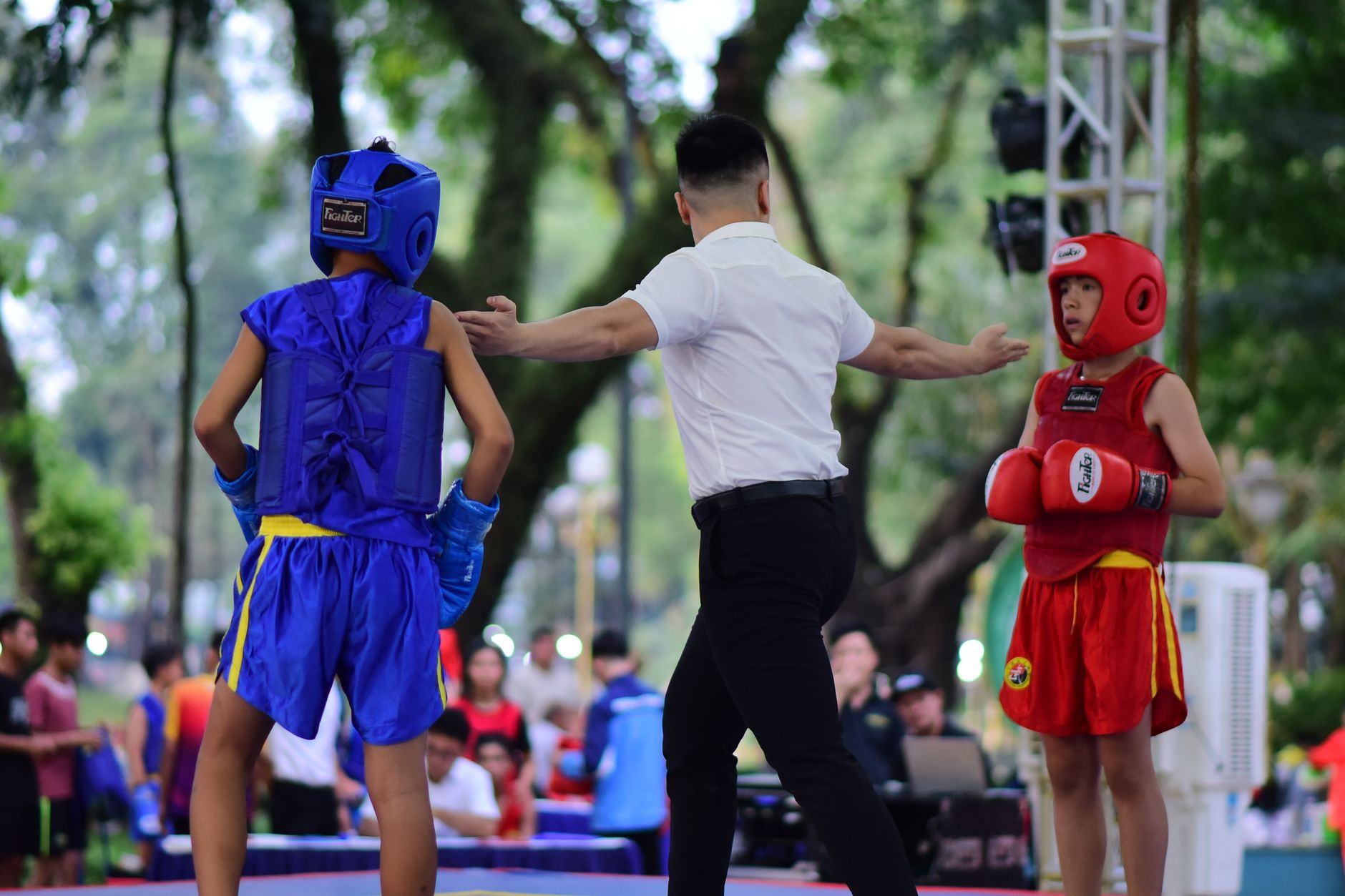 Two young boxers and a referee in an outdoor ring during a competitive match.