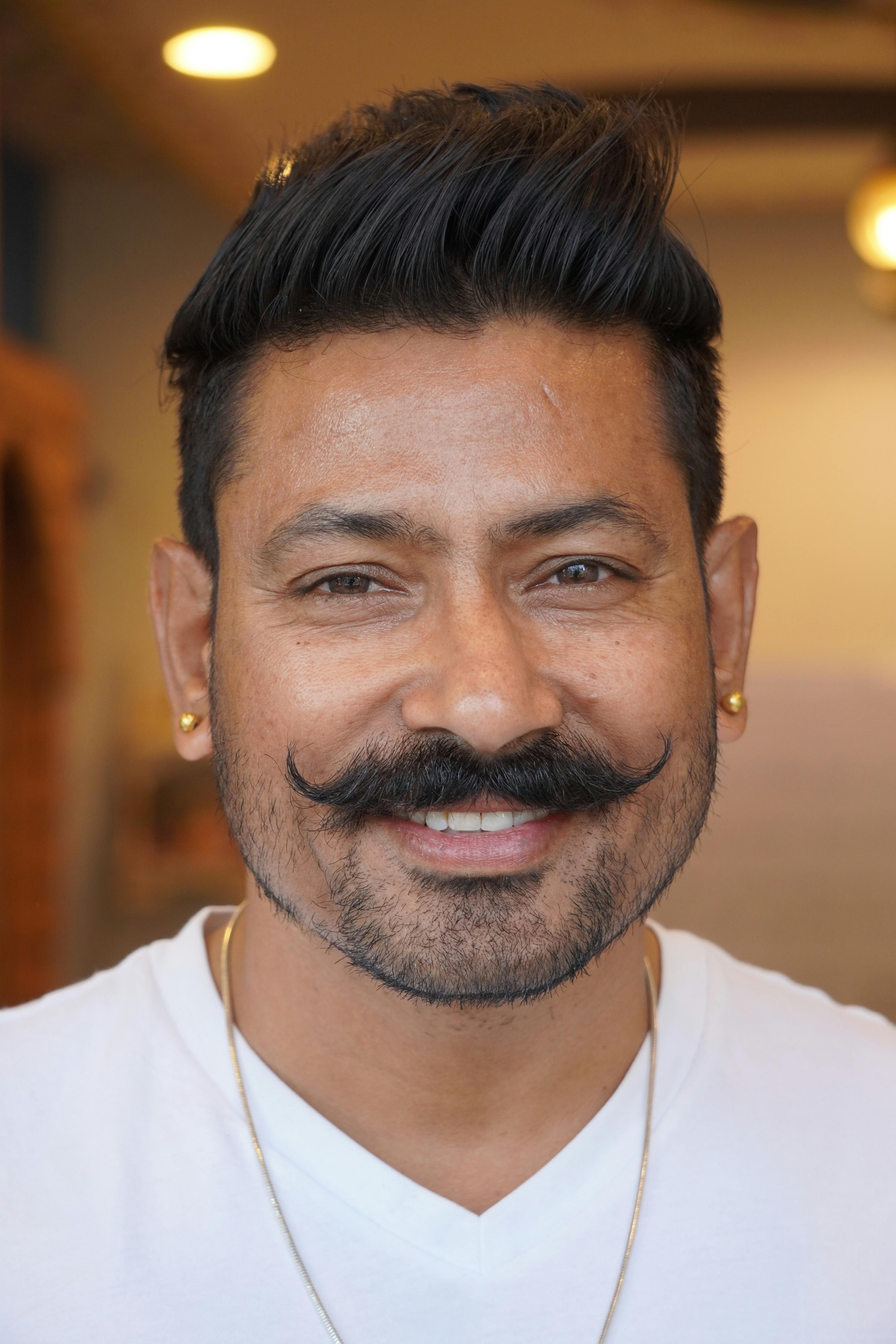 Close-up portrait of a smiling man with a styled mustache and earrings.