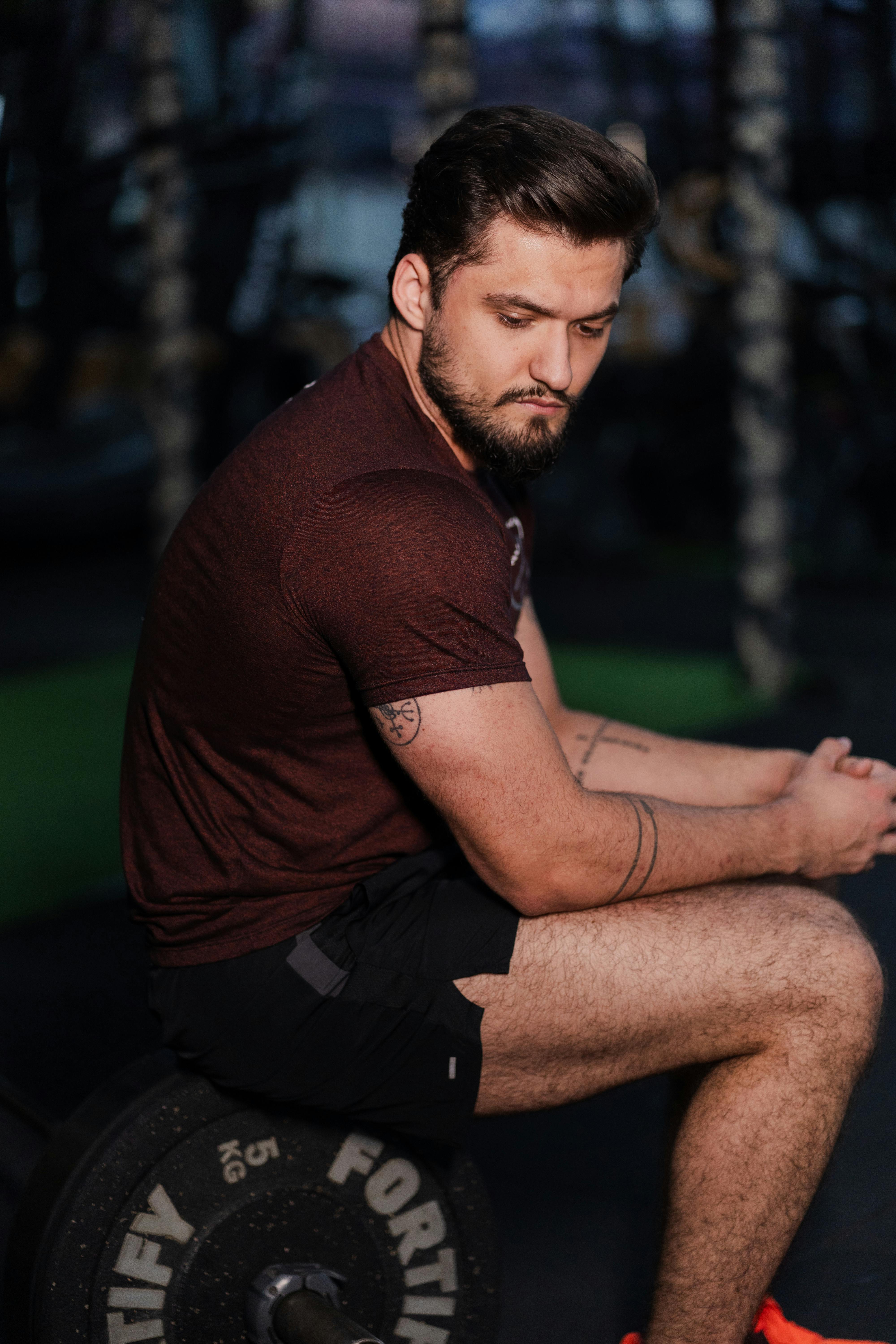 A muscular man sitting on a weight in a gym, reflecting between workout sets.